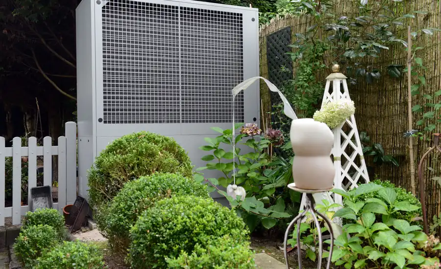 Außeneinheit einer Luft-Wasser-Wärmepumpe im Garten neben einem Zaun und Buchsbaumkugeln