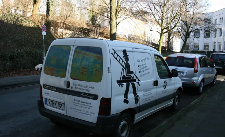 Weißer Kleintransporter mit Schornsteinfeger-Symbol und Beschriftung am Straßenrand vor Wohnhäusern