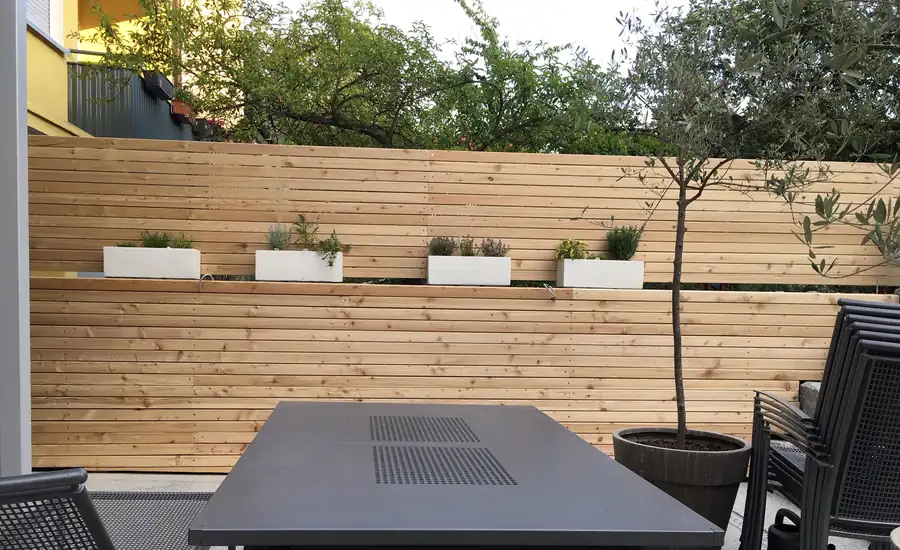 Mannshoher Sichtschutzzaun aus waagerechten Lärchenholzleisten mit Pflanzkästen an einer Terrasse mit Gartentisch, Stühlen und Olivenbäumchen