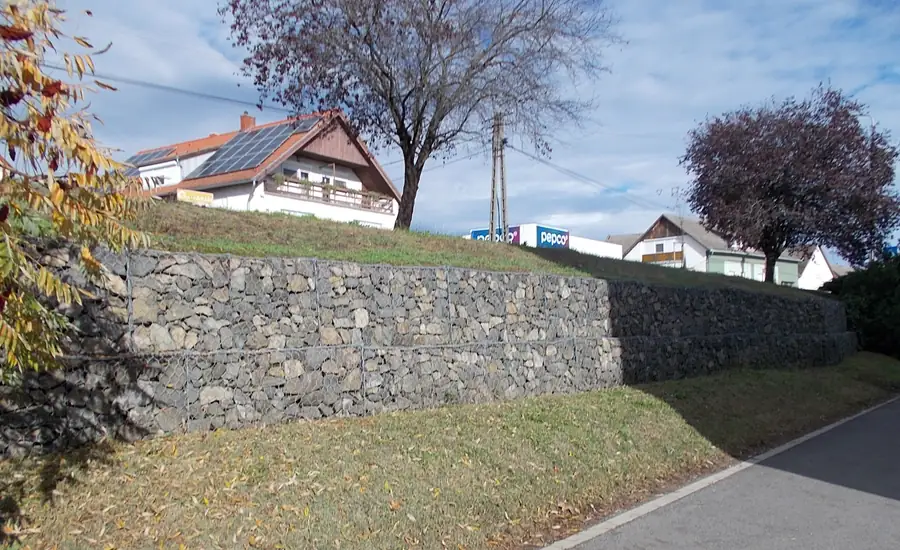 Gabionenwand aus Natursteinen entlang eines Gehwegs mit Böschung und Wohnhäusern im Hintergrund