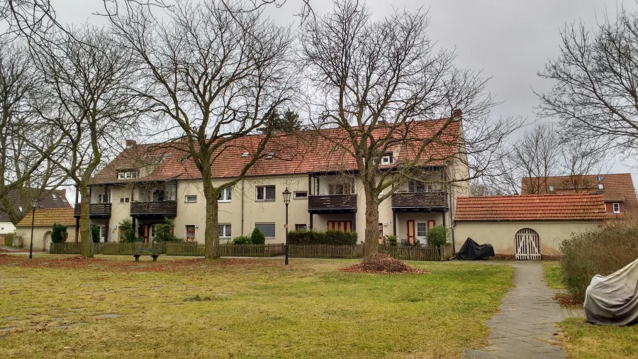 Zeile von Reihenhäusern mit rotem Satteldach und Holzbalkonen hinter einem Lattenzaun, davor eine Wiese mit kahlen Bäumen