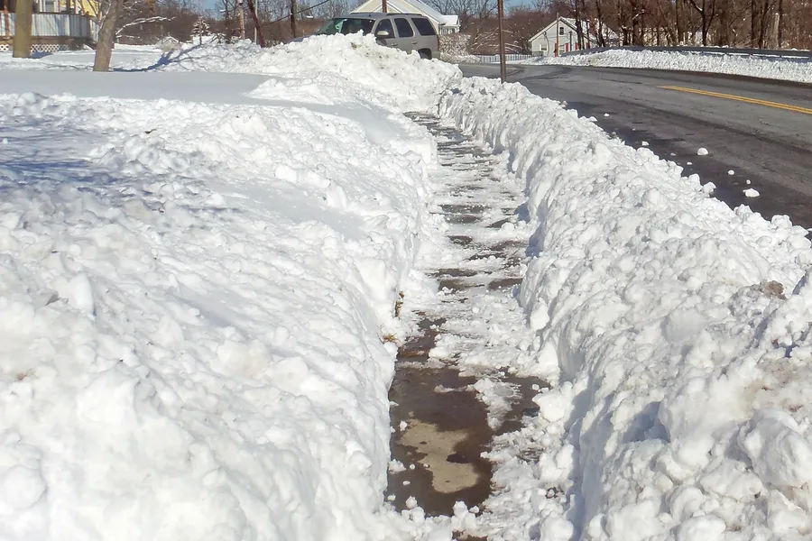 Mit der Schaufel freigeräumter schmaler Gehweg zwischen hohen Schneewällen neben einer Straße