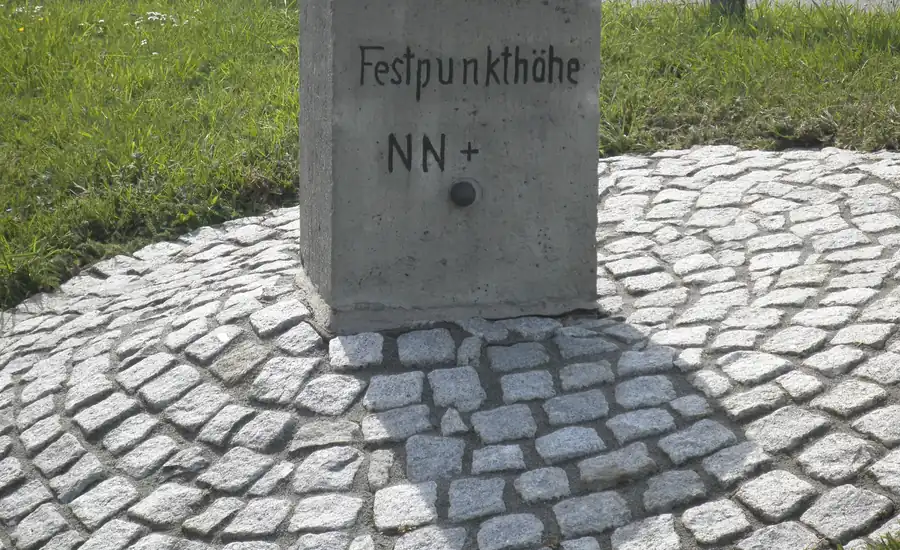 Steinerner Vermessungsfestpunkt mit der Aufschrift Festpunkthöhe NN und Höhenbolzen, eingefasst von Kopfsteinpflaster