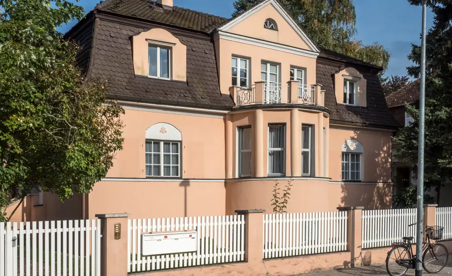 Rosa verputzte Villa mit Mansarddach hinter einem weißen Lattenzaun