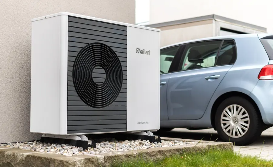 Außengerät einer Luft-Wärmepumpe auf einem Kiesstreifen neben einer Hauswand, dahinter ein geparktes Auto