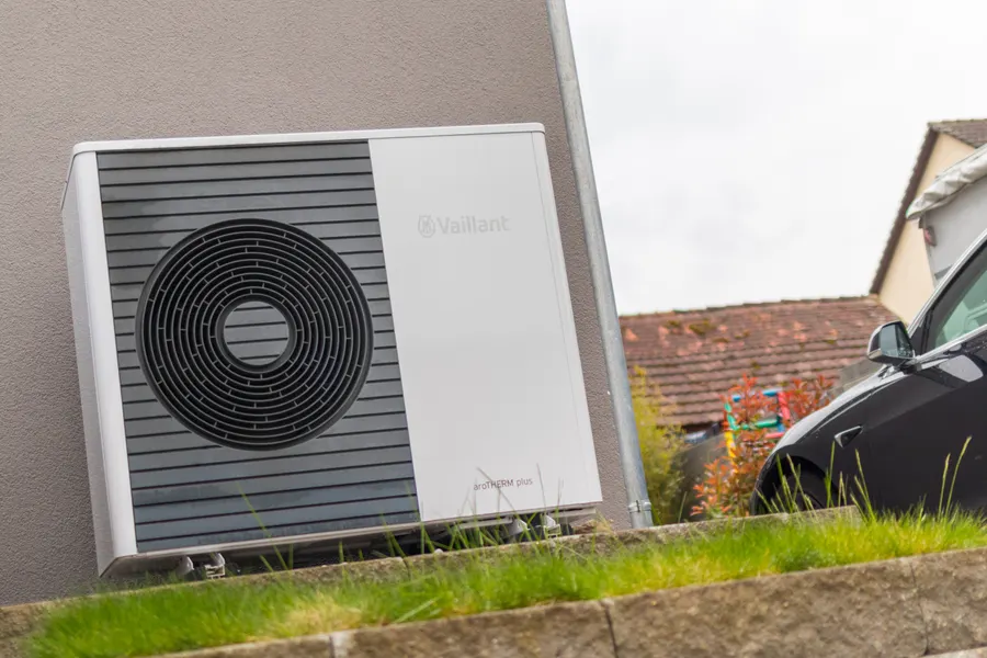 Symbolfoto: Außeneinheit einer Luft-Wasser-Wärmepumpe an der Wand eines Hauses, davor ein Kiesbett und ein Mauerrand, rechts ein geparktes Auto