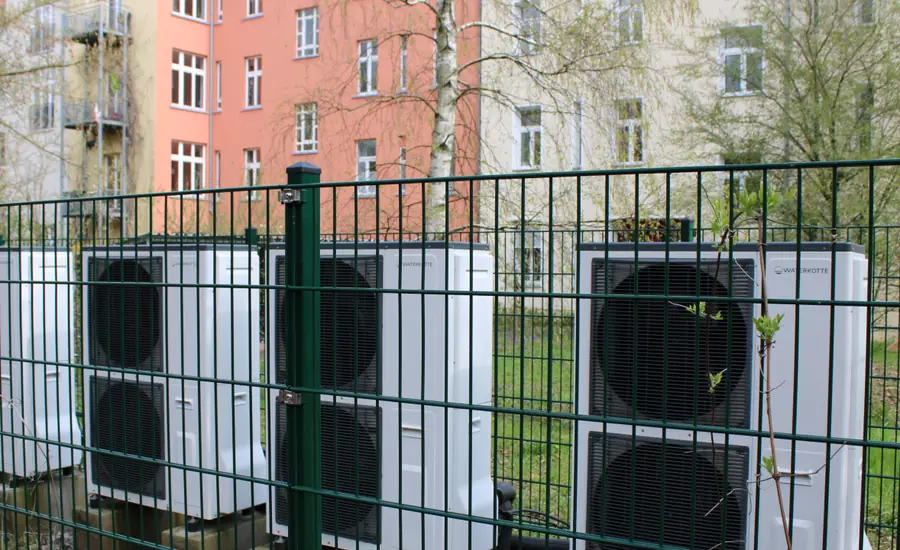 Drei Wärmepumpen-Außengeräte hinter einem Zaun an der Grundstücksgrenze, dahinter ein Wohngebäude