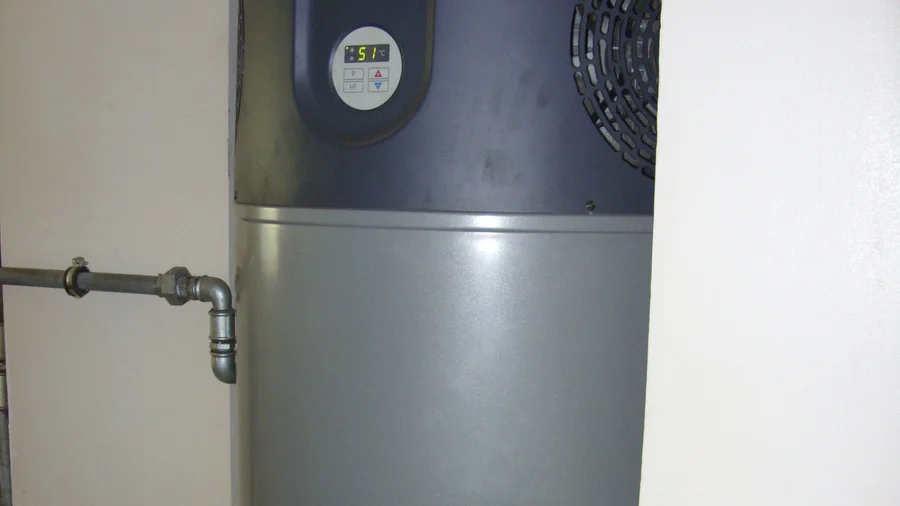 Grauer Warmwasserspeicher mit Wärmepumpenaufsatz, Lüftungsgitter, Temperaturanzeige und angeschlossenem Rohr