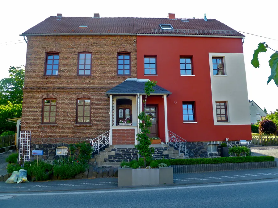 Zweigeschossiges Haus mit Satteldach: links Backsteinfassade, rechts rot und beige verputzte Fassade mit tiefen Fensterlaibungen