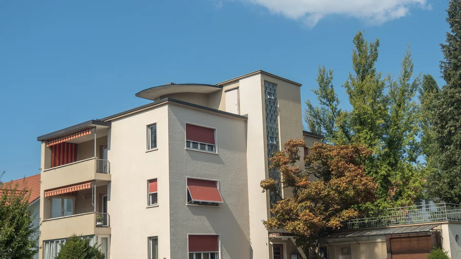 Mehrgeschossiges Wohnhaus mit Balkonen, roten Rollläden und Bäumen