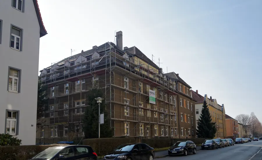 Mehrfamilienhaus mit Baugerüst am oberen Geschoss, davor parkende Autos an der Straße