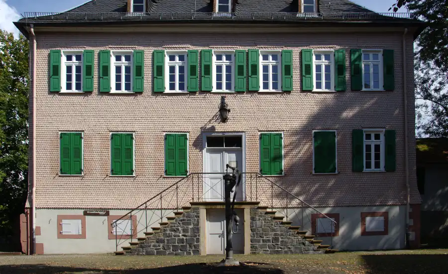 Mit Holzschindeln verkleideter Bau mit grünen Fensterläden, doppelläufiger Steintreppe und alter Schwengelpumpe davor: das Schloss in Nidda, in dem das Grundbuchamt sitzt.