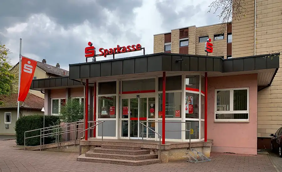 Eingang einer Sparkassen-Filiale in Wertheim-Bestenheid mit rotem Logo über dem verglasten Vorbau, Treppe mit Geländer und rotem Sparkassen-Banner.